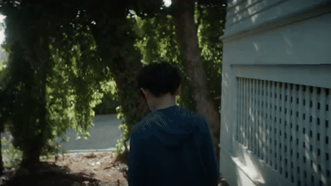 Boy walks along the side of a house and a fence in the tree lined yard. From Alex Cook's film "Undertow" by Son Lux.